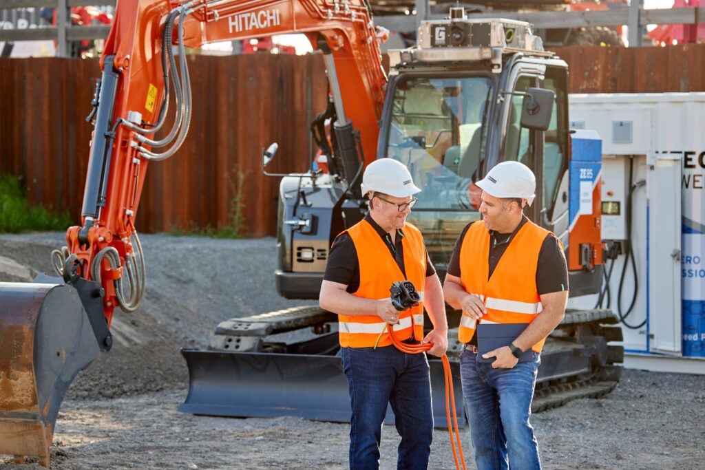 Two men in safety vests and helmets talking on a construction site in front of an electric Hitachi excavator.