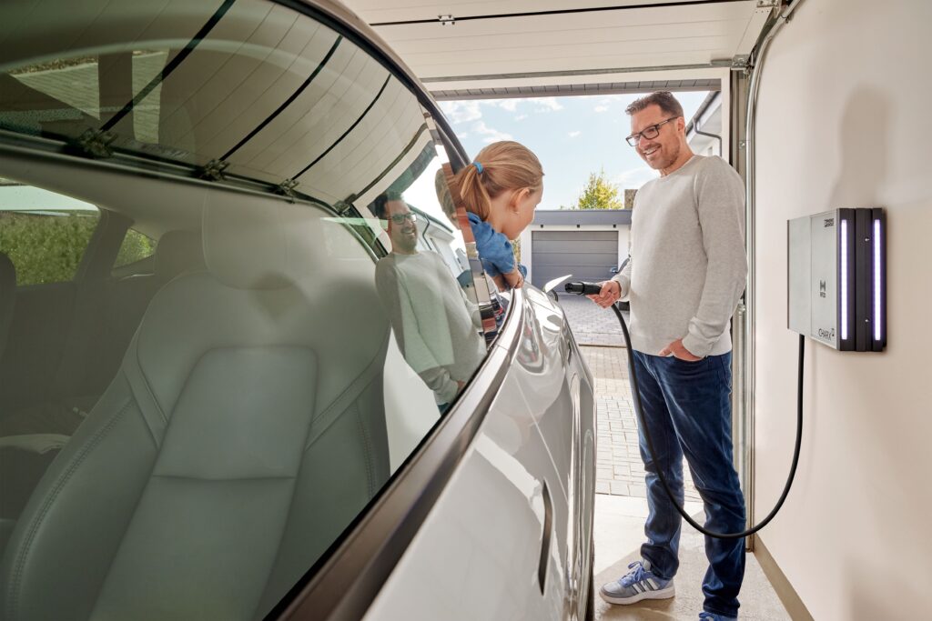 Mann lädt in seiner Garage ein Elektroauto mit einer Wallbox von CHARX, während ein Kind neugierig durch das geöffnete Fenster schaut.