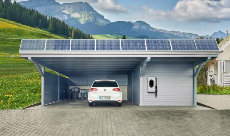 A modern carport with a solar panel on the roof stands in front of a picturesque mountain landscape. A white electric car is parked under the carport, connected to a wall-mounted charging station. Green meadows, trees and mountains can be seen in the background, the sky is partly cloudy.