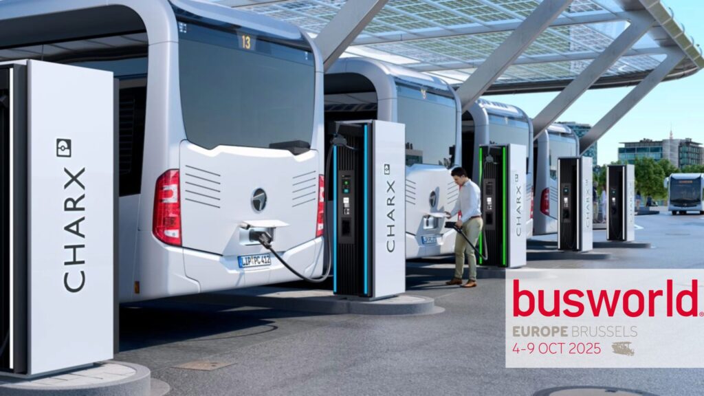 A row of electric buses is being charged at CHARX charging stations. A person is connecting one of the buses to a station. In the background, a modern bus terminal with solar panels on the roof is visible. Text in the image reads: ‘busworld EUROPE BRUSSELS 4–9 OCT 2025.