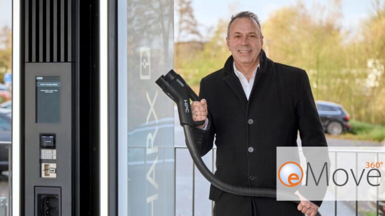 Joachim Pucker, Director Sales E-mobility, stands in front of a charging station for electric vehicles and holds an HPC charging cable. Trees and parked cars can be seen in the background. The eMove360° logo is visible on the right of the picture.