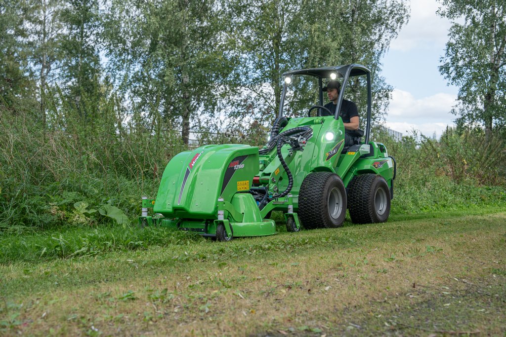 Eine Person bedient einen grünen Avant-Lader mit einem Mähwerk in einem von Bäumen und Grünflächen umgebenen Außenbereich. Der Lader mäht Gras unter einem teilweise bewölkten Himmel in einer natürlichen Umgebung.