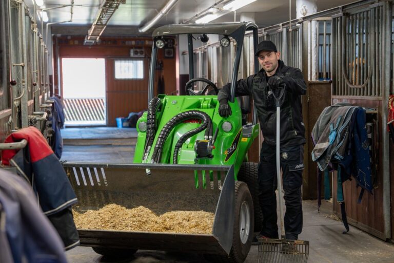 A person stands next to a green Avant compact loader inside a horse stable, holding a pitchfork.