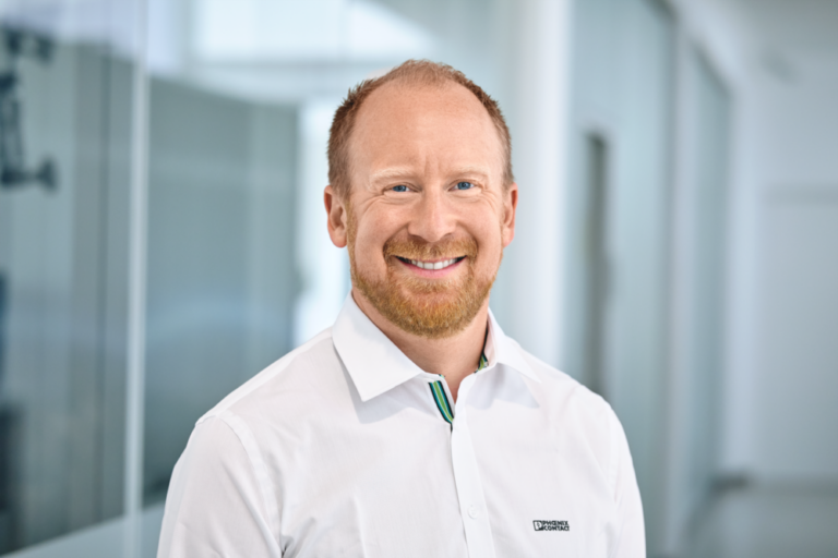 Person in einem weißen Hemd mit Phoenix Contact-Logo steht in einem modernen Büroflur mit Glaswänden und heller Beleuchtung.