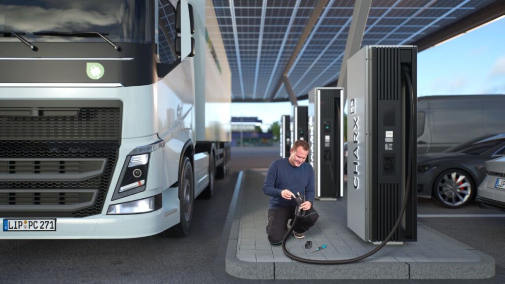 Person arbeitet an einer Ladeeinrichtung für Elektrofahrzeuge unter einem überdachten Ladepark mit Solarpanel-Dach. Links steht ein großer Lkw, rechts ein Pkw, und im Vordergrund ist eine CHARX-Ladesäule mit Kabel sichtbar.
