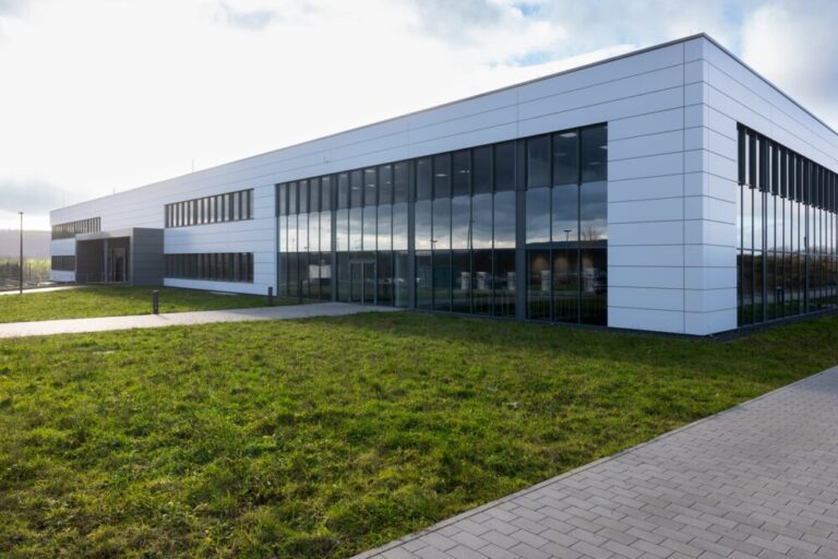 Modernes Industriegebäude mit weißer Fassade und großen Glasfronten, umgeben von einer Grünfläche und einem gepflasterten Gehweg, unter leicht bewölktem Himmel.