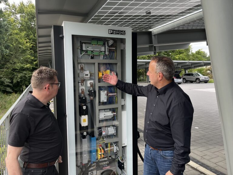 Two men stand in front of an outdoor DC charging station. It bears the logo 'PHOENIX CONTACT' and contains electronic components, cables and circuits. One of the men points to a component inside while the other watches intently. Trees and a parking lot with cars can be seen in the background.